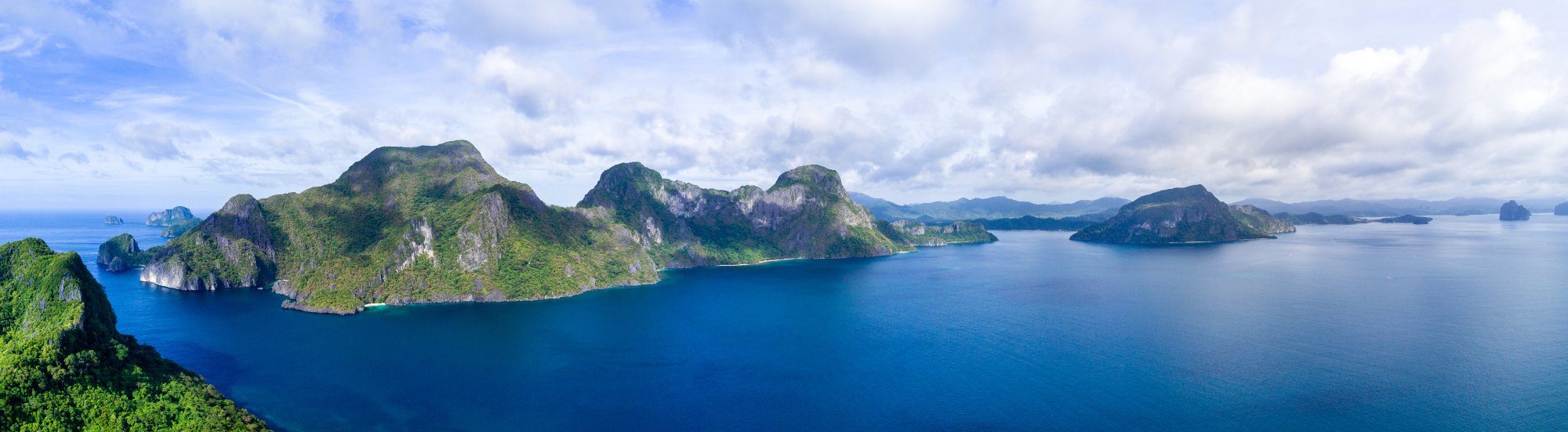 A stunning aerial view of a lush tropical archipelago featuring steep limestone cliffs covered in dense green vegetation, rising dramatically from crystal-clear turquoise waters. Multiple islands of varying sizes are scattered across the seascape under a partly cloudy sky, suggesting a Southeast Asian destination popular with cruise itineraries.