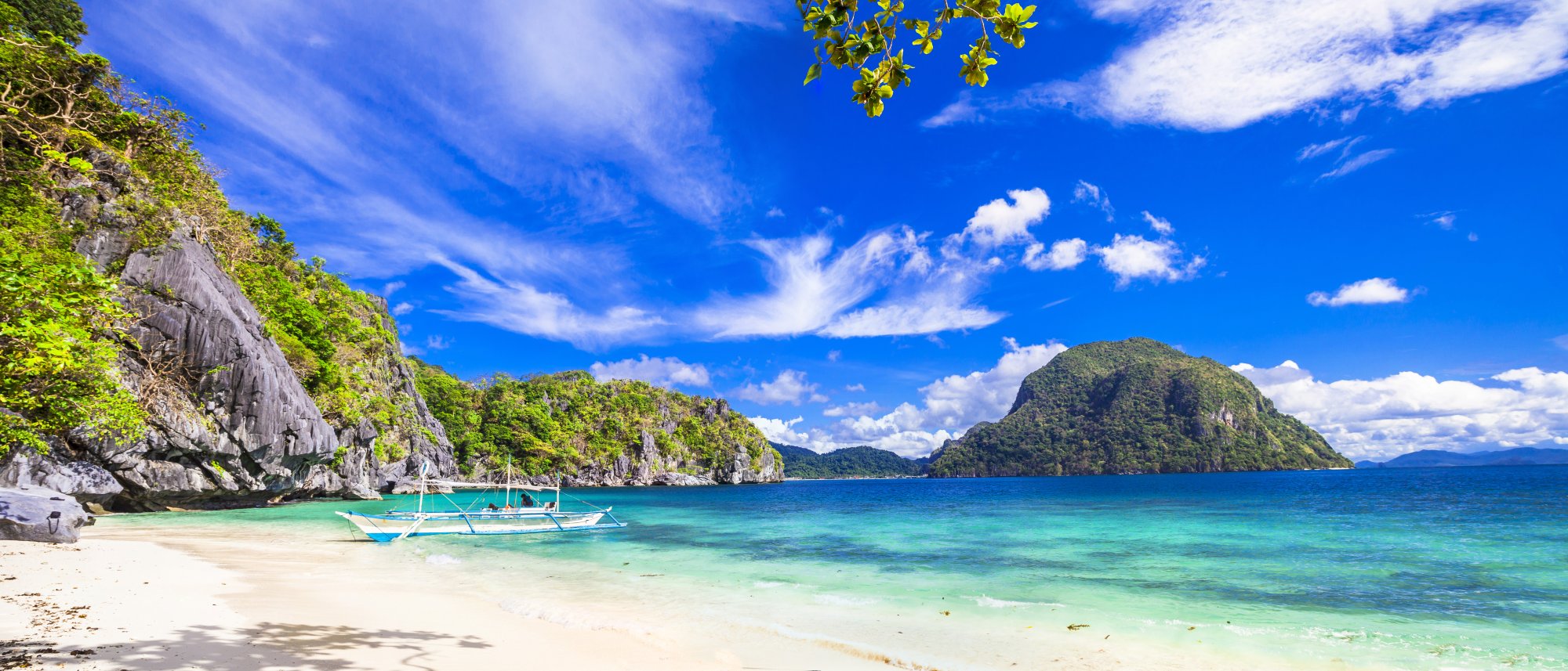 A pristine tropical beach nestled beneath towering limestone cliffs, featuring crystal-clear turquoise waters, white sand, and traditional outrigger boats anchored offshore. Lush green vegetation covers the dramatic rock formations, with a distant volcanic island visible across the bay under a bright blue sky.