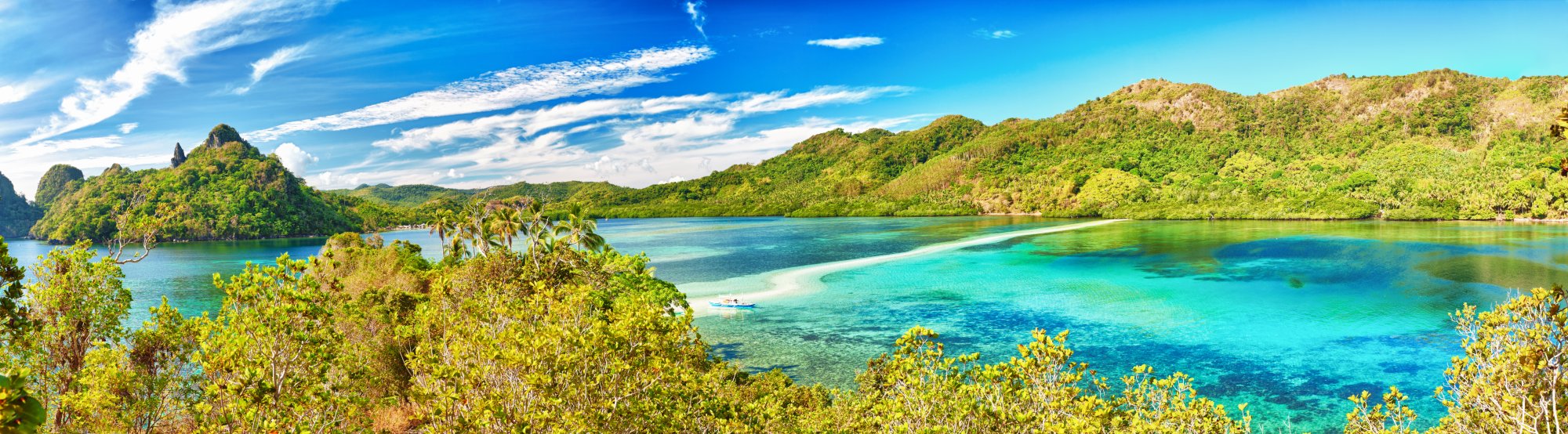A stunning aerial view of a pristine tropical lagoon featuring crystal-clear turquoise waters, white sand beaches, lush green mangrove forests, and dramatic limestone cliffs under a bright blue sky. The scenic landscape captures the natural beauty of a remote island destination ideal for cruise tourism and water activities.