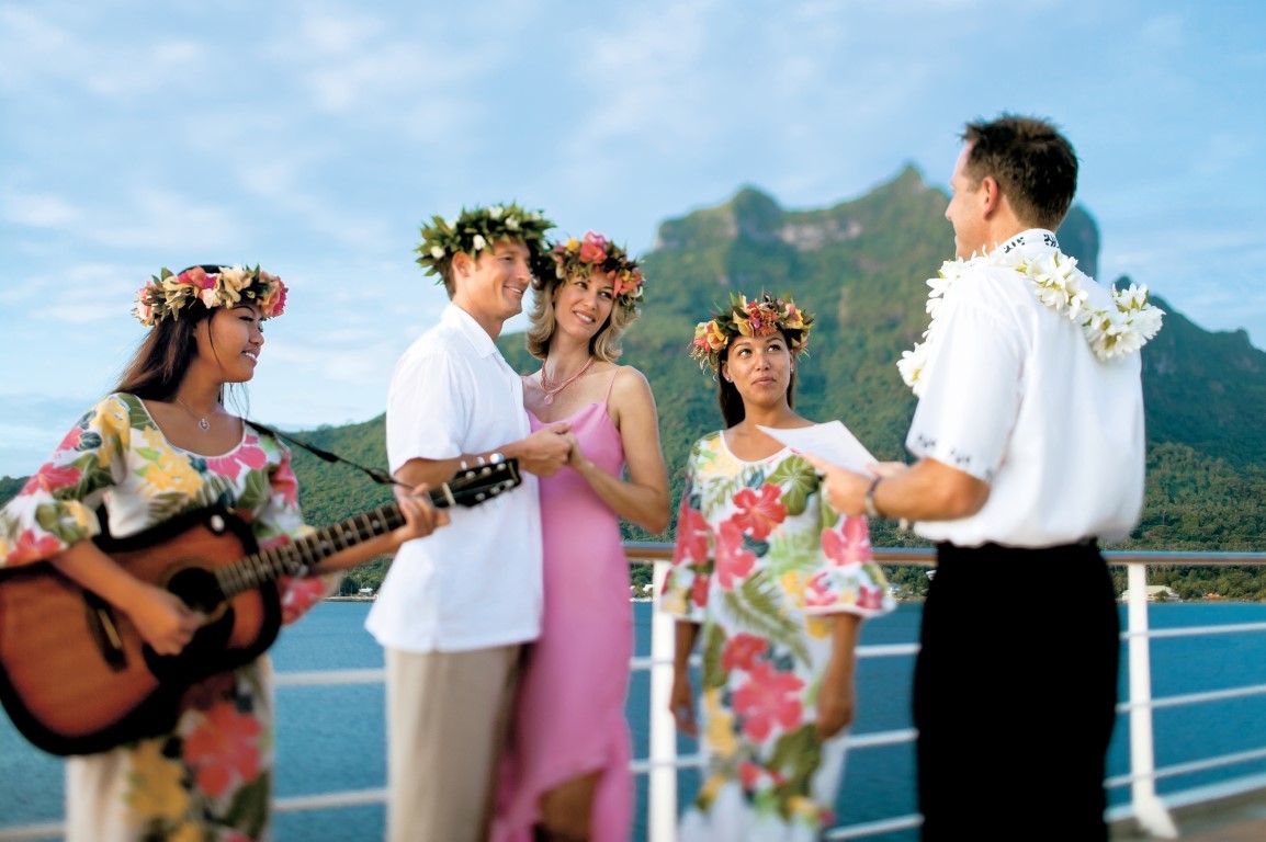 A joyful wedding celebration on a cruise ship deck featuring five people in tropical attire and flower garlands, with a lush green volcanic island and turquoise ocean visible in the background.