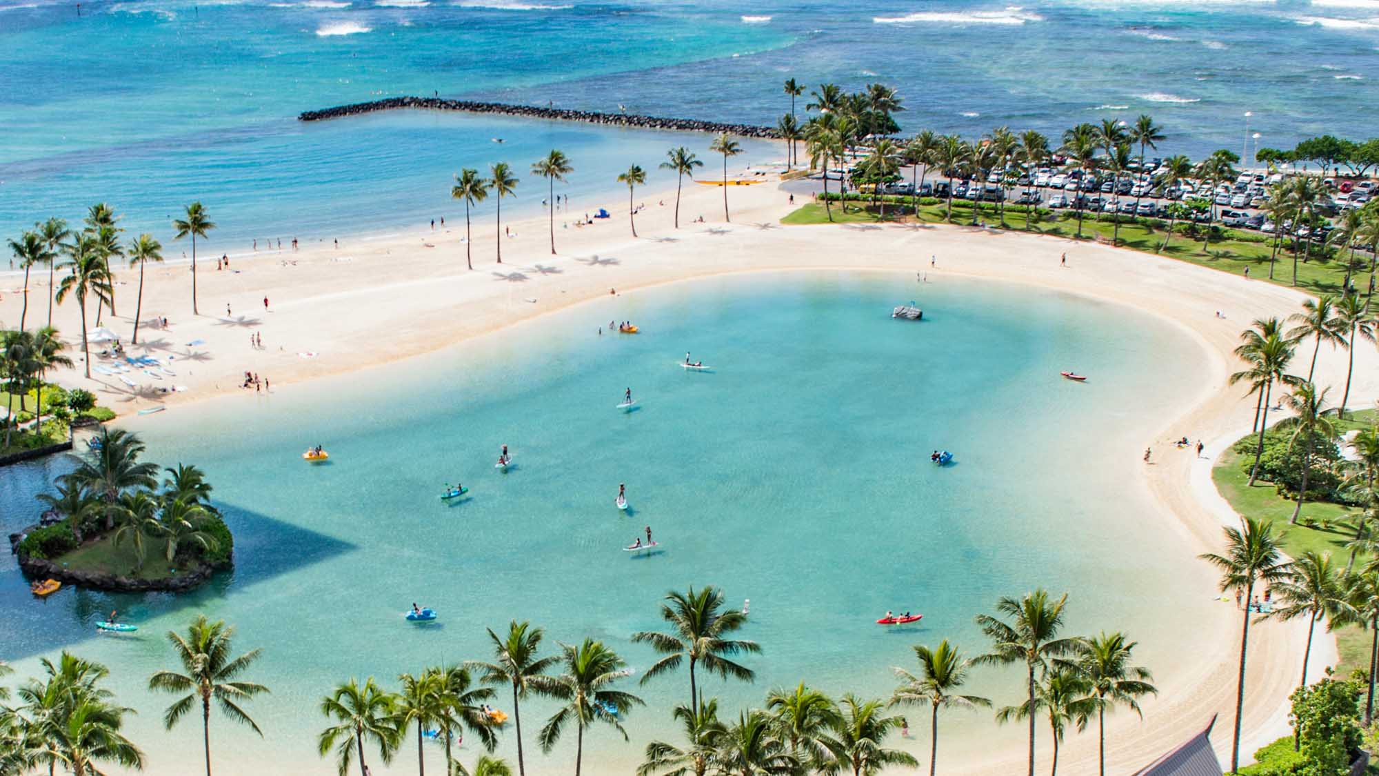 Tropical Paradise Beach with Palm Trees and Turquoise Lagoon An aerial view of a pristine tropical beach destination featuring a stunning turquoise lagoon with soft white sand, scattered palm trees, and numerous water activities. The scene includes a parking area, grassy park space, and a protective breakwater, showcasing a popular beach resort destination with calm, crystal-clear waters ideal for swimming and water sports.