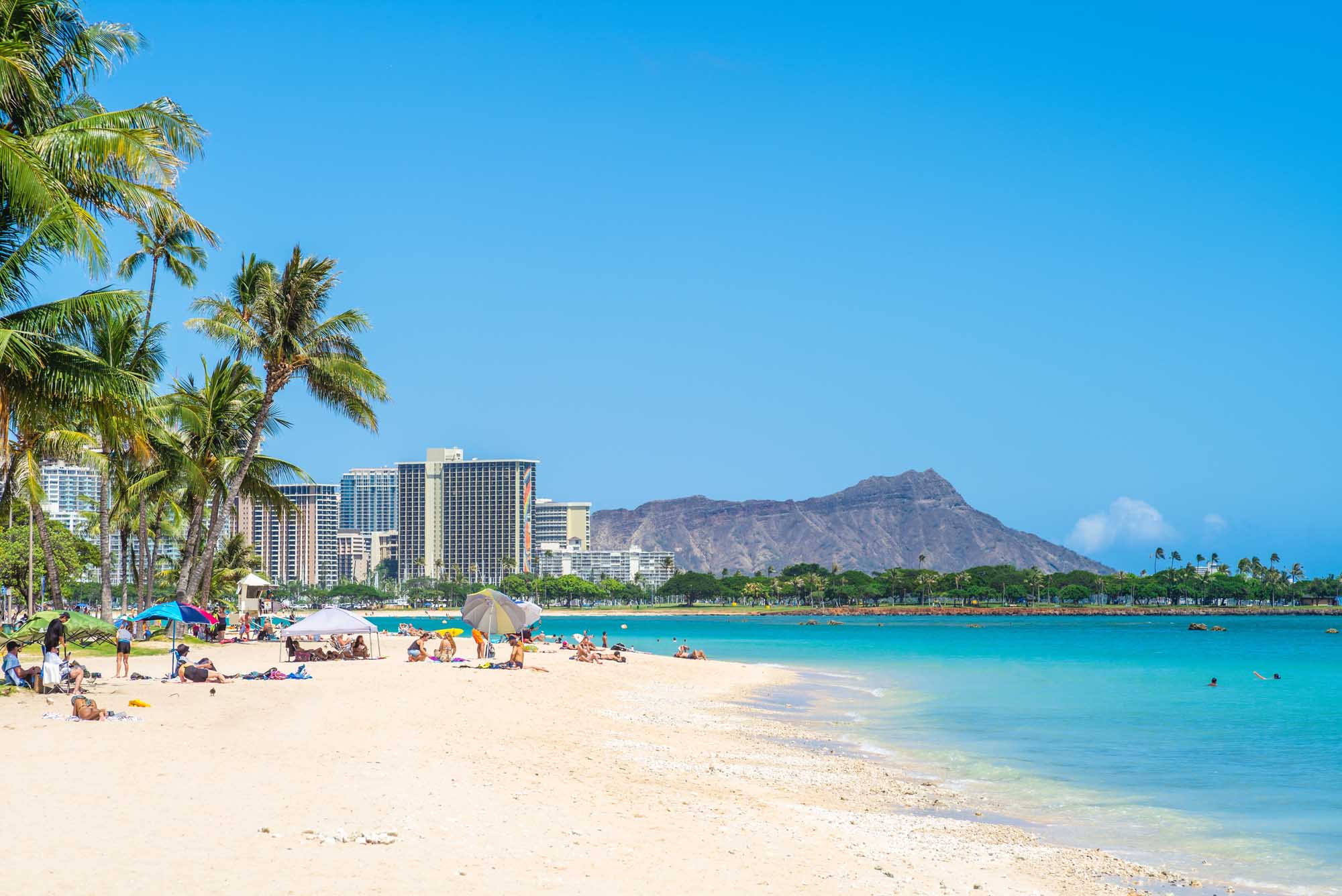 Waikiki Beach with Diamond Head Mountain A scenic view of Waikiki Beach in Honolulu, Hawaii, featuring pristine white sand, crystal-clear turquoise waters, and beachgoers enjoying the tropical weather. The iconic Diamond Head mountain rises majestically in the background, with modern high-rise hotels and swaying palm trees framing this quintessential Hawaiian destination.