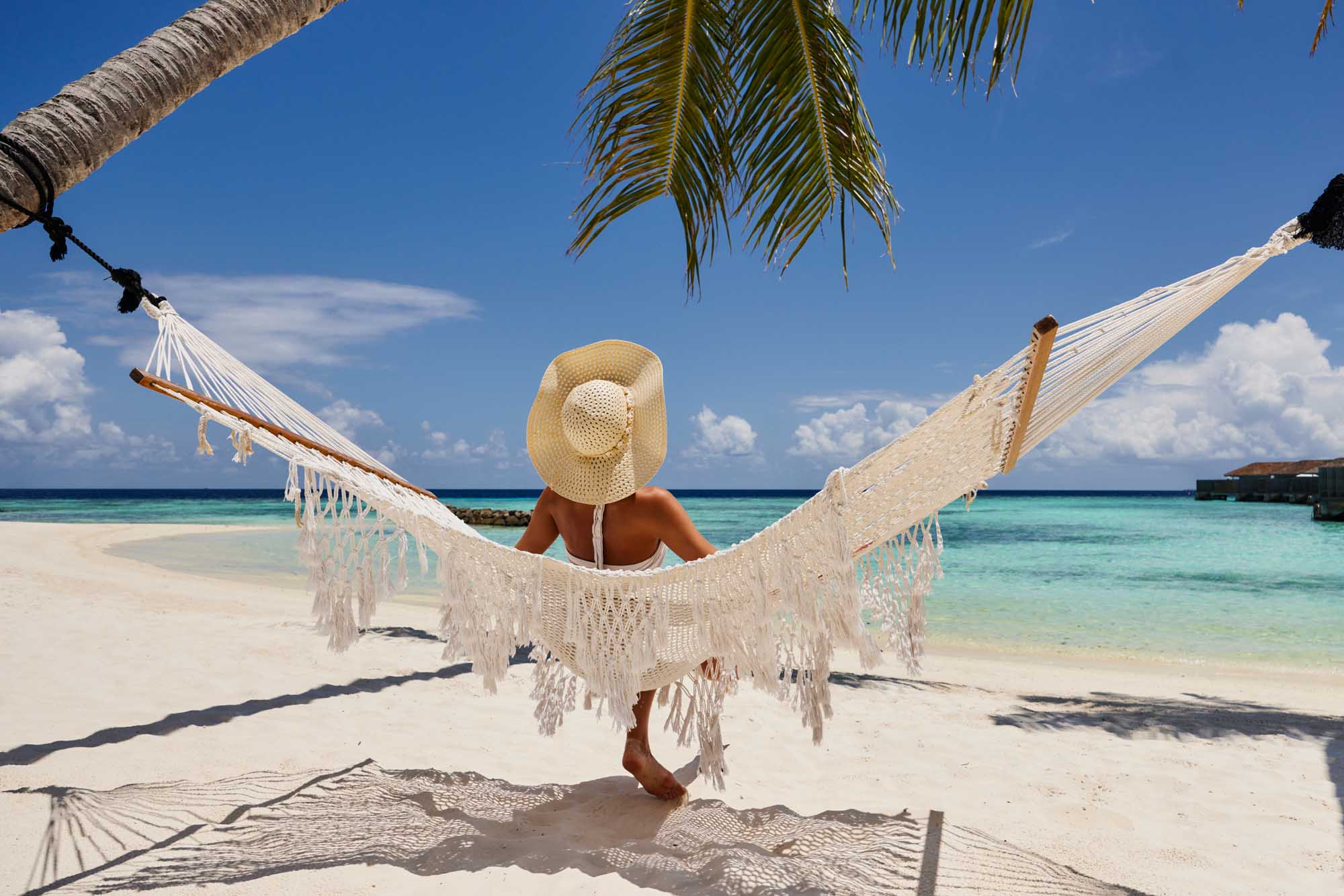 Woman Relaxing in Hammock on Tropical Beach A rear view of a woman wearing a straw sun hat relaxing in a white macramé hammock strung between palm trees on a pristine tropical beach, overlooking turquoise waters and a clear blue sky dotted with white clouds.