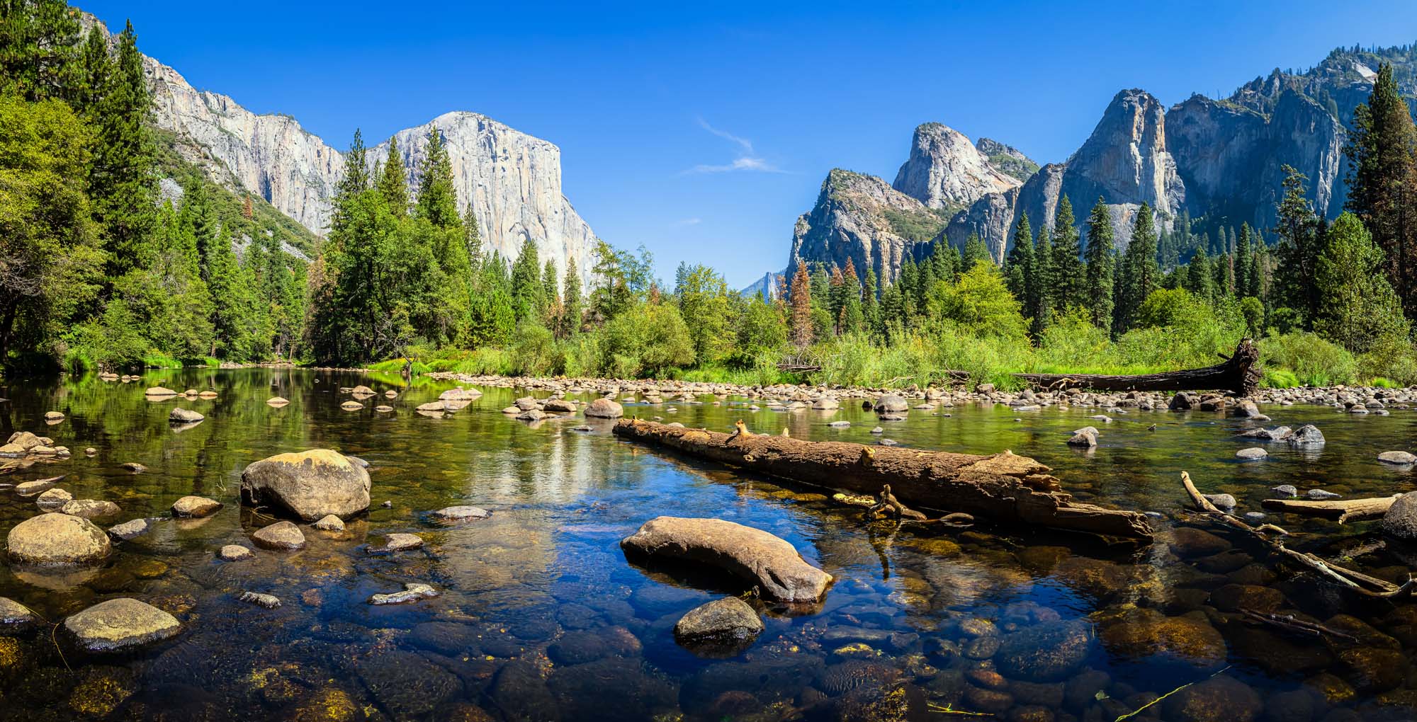A stunning panoramic view of Yosemite Valley featuring the iconic granite cliffs of El Capitan and Three Brothers formations rising majestically above a pristine stretch of the Merced River. The scene captures clear reflections in the calm water with scattered boulders, dense coniferous forest, and a brilliant blue summer sky.