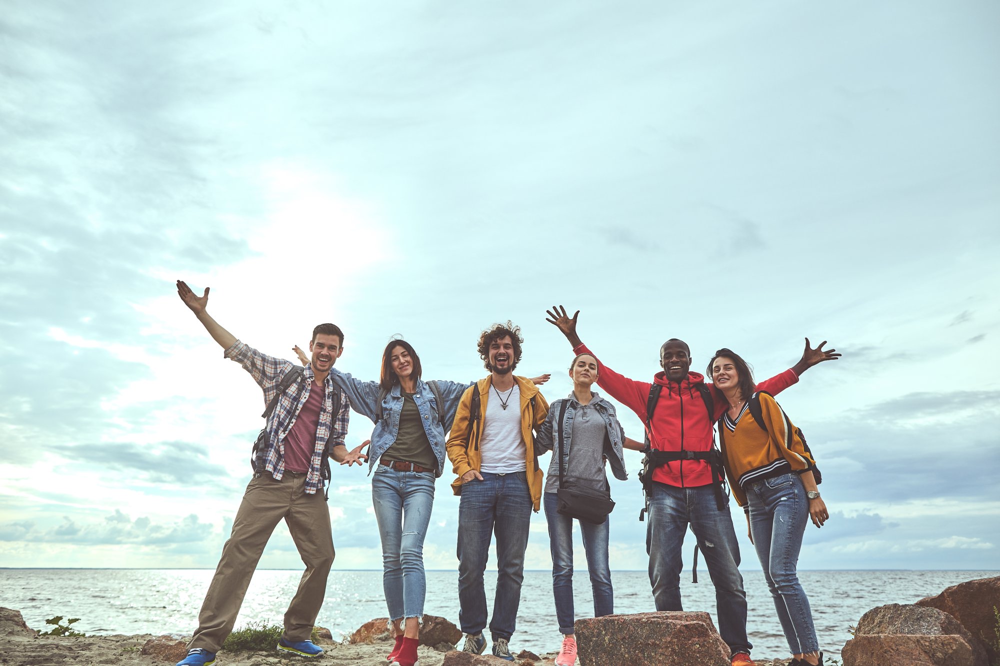 A diverse group of six young adults standing on rocks by the ocean with their arms raised in celebration and joy, capturing a moment of happiness and camaraderie during a travel outing.