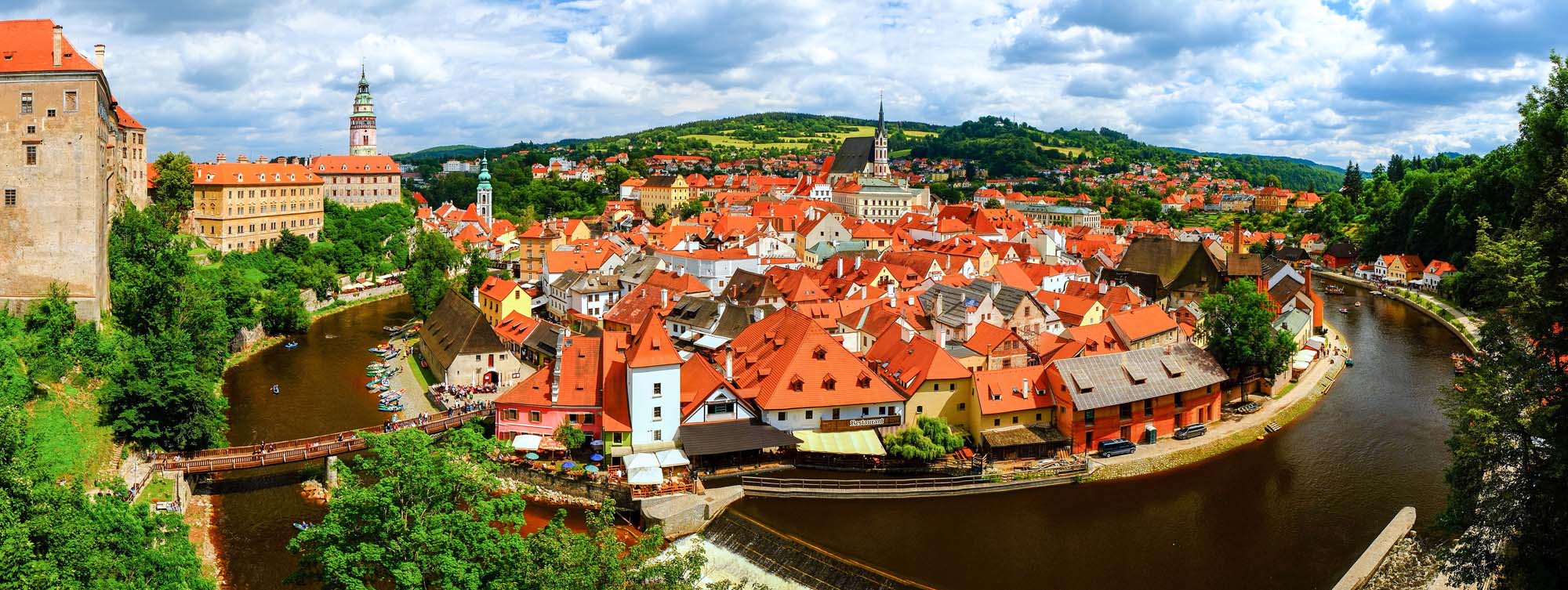 A stunning panoramic view of Český Krumlov, a UNESCO World Heritage Site in the Czech Republic, featuring the iconic red-tiled roofs of the medieval old town nestled within a dramatic bend of the Vltava River, with the historic castle and church spires rising prominently against forested hills under a partly cloudy sky.