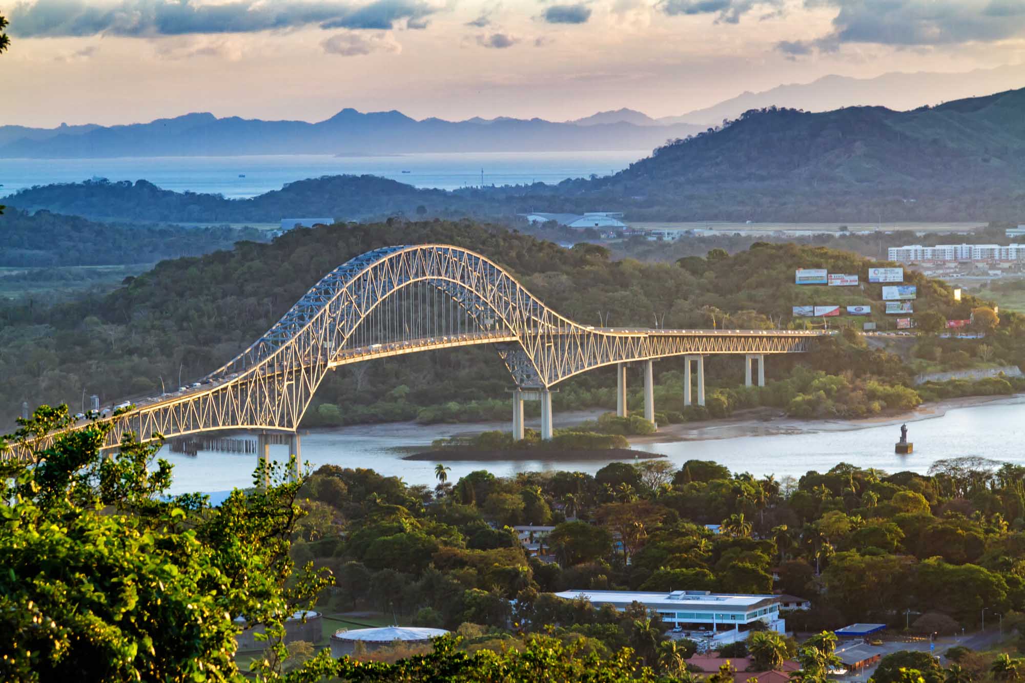 A stunning arched bridge with golden-yellow steel lattice construction spans a wide river in a lush, mountainous tropical landscape with dense green forests and distant mountains across a bay.