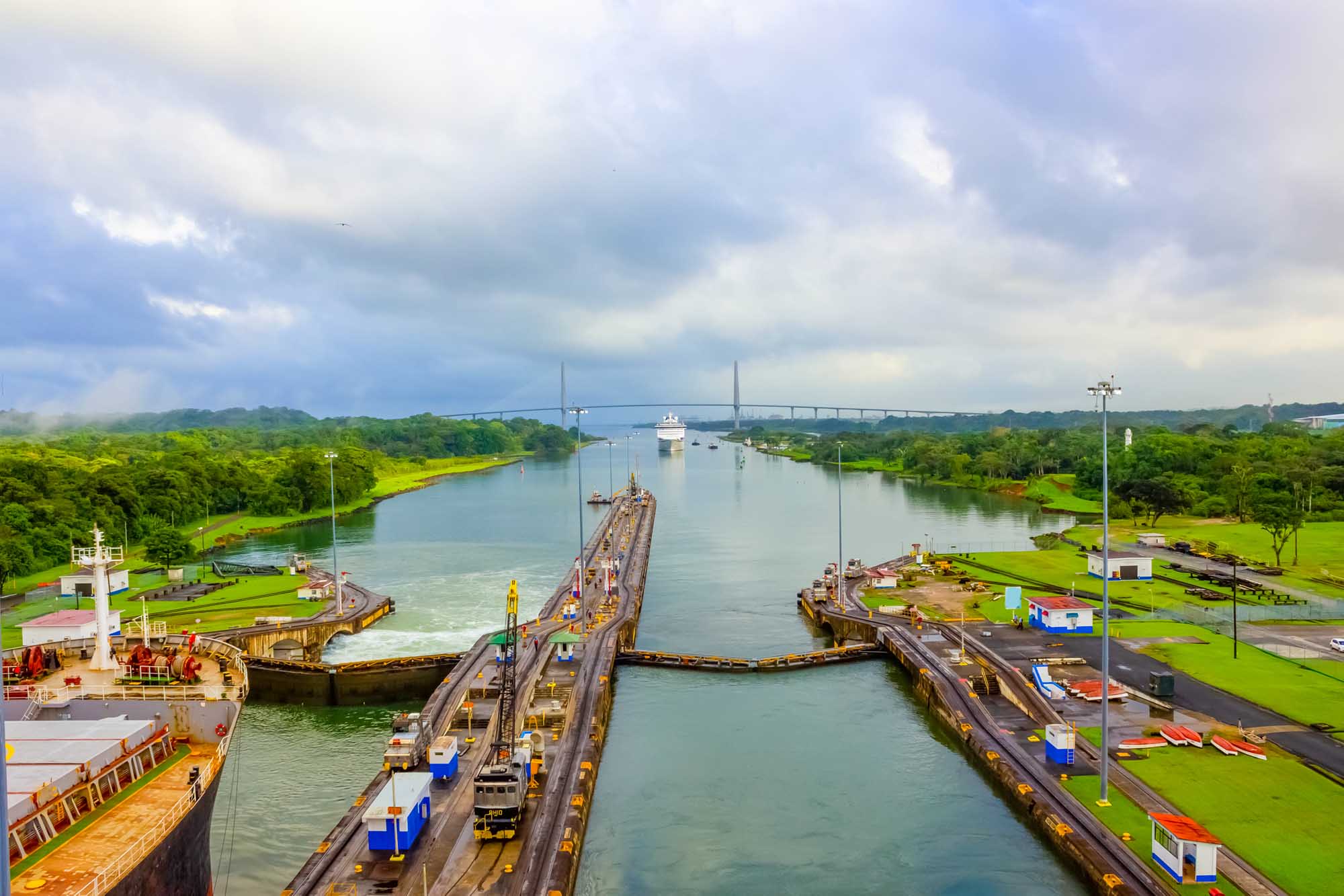 An aerial view of the Panama Canal's lock system showing massive lock chambers with vessels in transit, with a bridge spanning the canal in the background and lush tropical landscape surrounding the infrastructure.