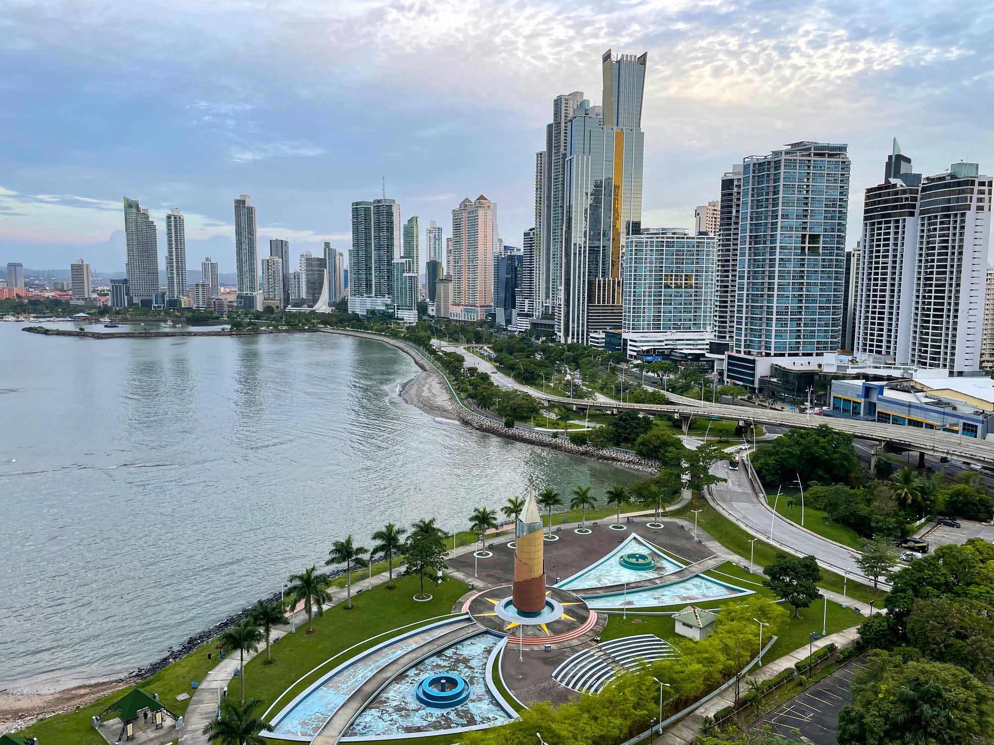 A panoramic view of a modern high-rise skyline stretching along a curved sandy beach, with lush green parklands and a distinctive sculptural monument in the foreground.