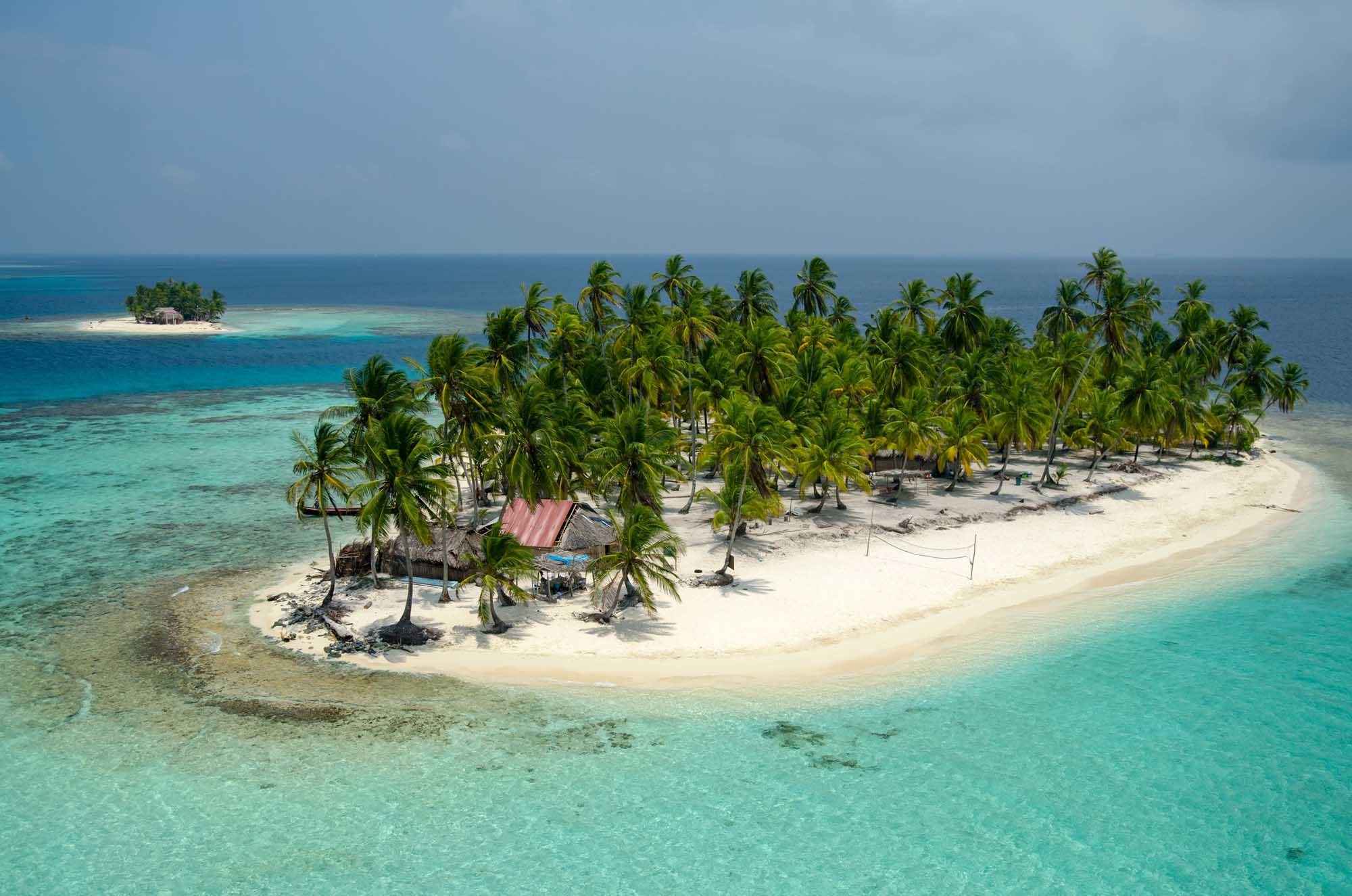 An aerial view of a picturesque tropical atoll featuring a narrow strip of white sand beach lined with coconut palm trees, surrounded by stunning turquoise and azure waters.