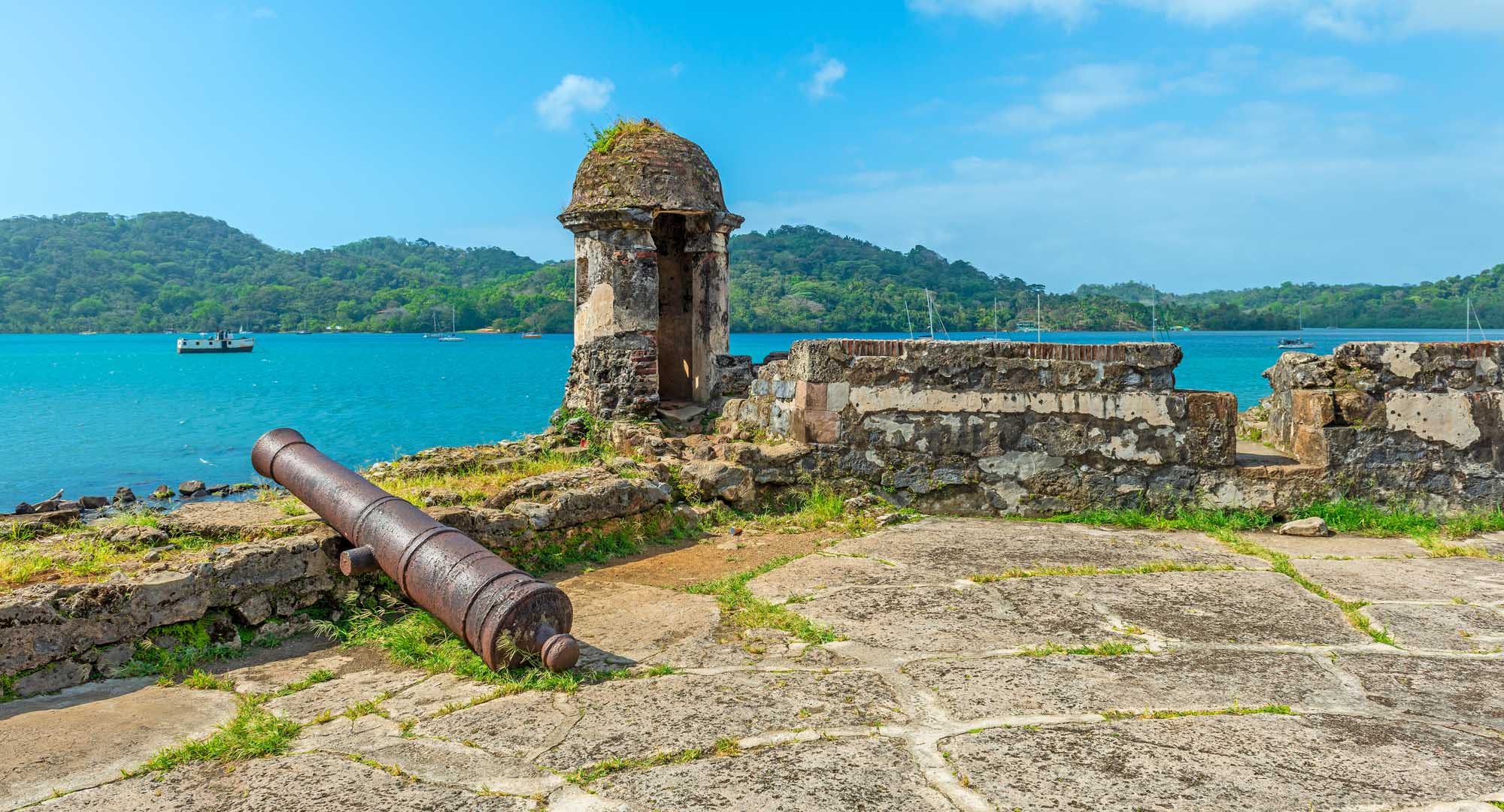 Weathered stone fortress ruins with a distinctive domed sentinel tower and corroded cannon overlook a stunning turquoise bay dotted with sailing vessels and lush forested hills.