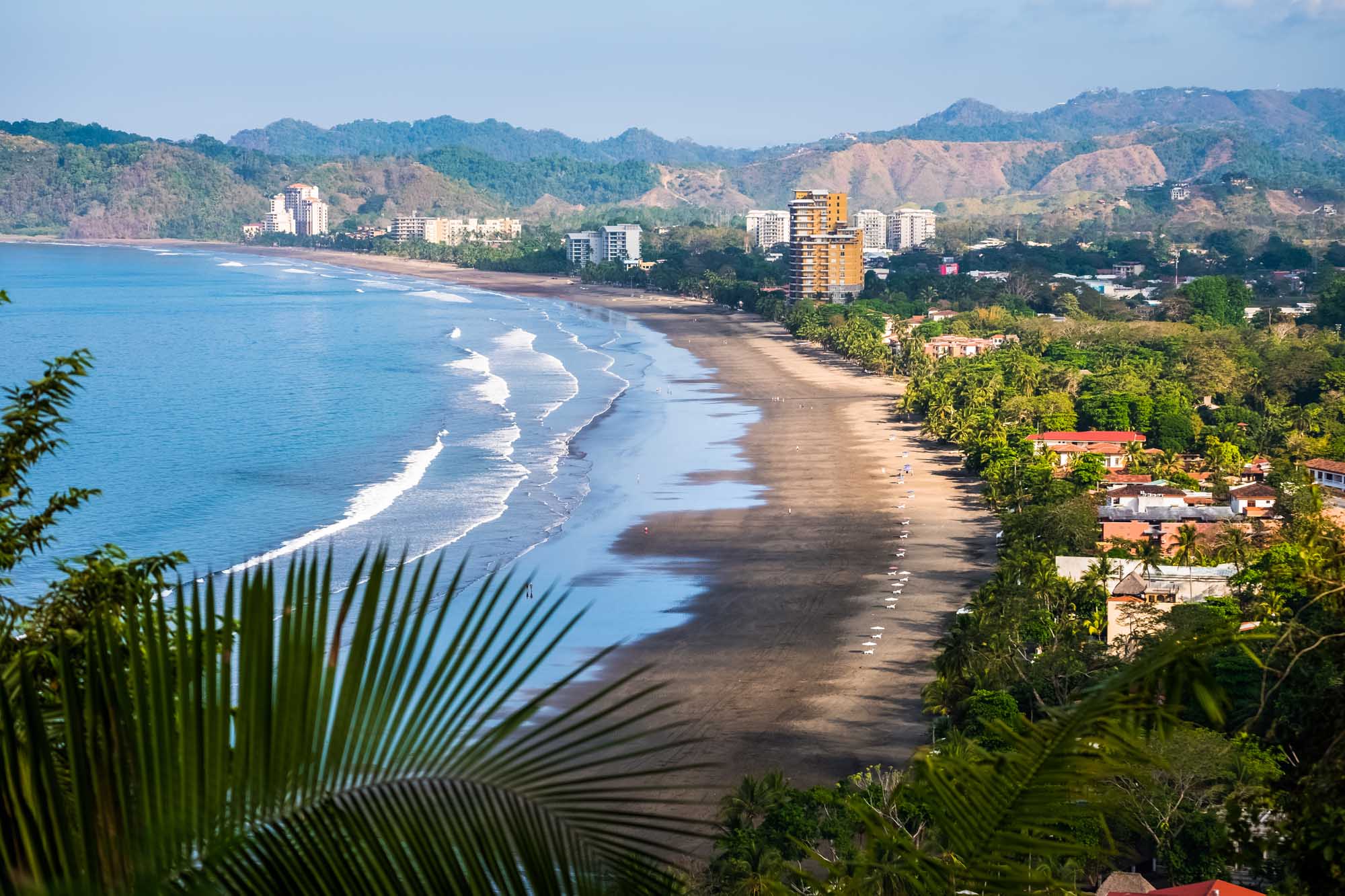 A scenic coastal vista featuring a pristine beach with gentle waves, flanked by lush tropical vegetation and a modern beachfront resort town nestled against verdant, forested mountains.