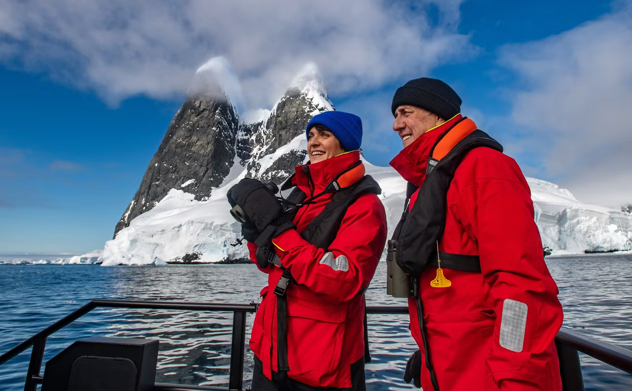 Expedition Couple Observing Antarctic Landscape Two expedition-dressed travelers in bright red parkas stand on the deck of a zodiac or small