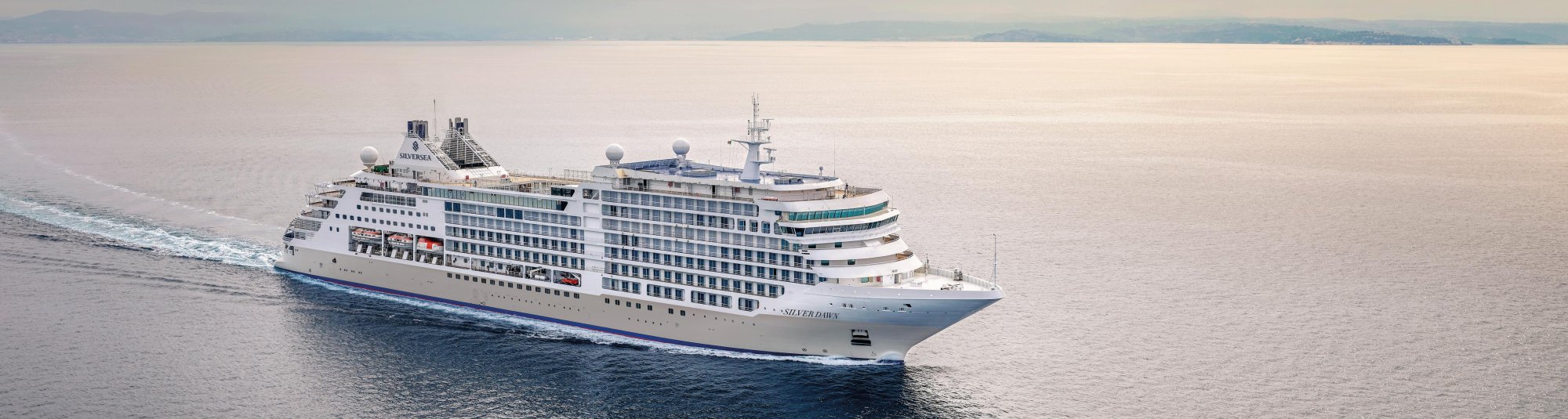 A large luxury cruise ship from Silversea navigates through calm waters with distant mountains