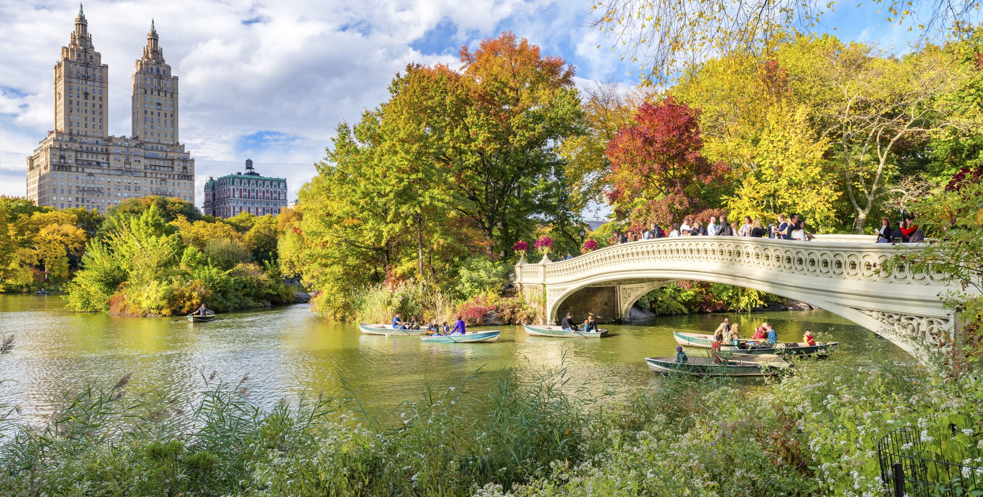 A scenic autumn view of Bow Bridge spanning a serene lake in Central Park, with vibrant fall