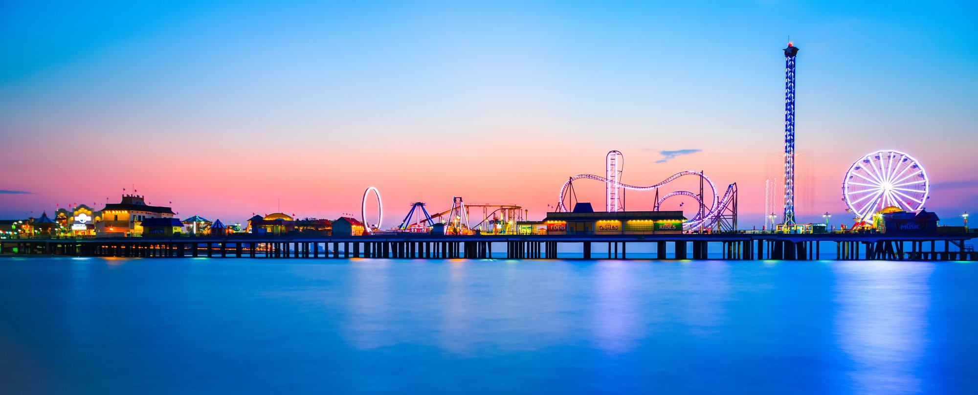 An illuminated amusement pier extending into calm blue waters at dusk, featuring a Ferris wheel